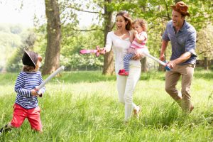 Laughing mother holds baby while father and other child duel with toy swords in grassy field