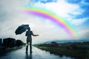 A person in business wear holds up umbrella with hand out to check for rain, looking at a rainbow in a cloudy sky