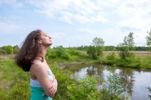 Person with long curly hair faces out over lake surrounded by trees with eyes closed