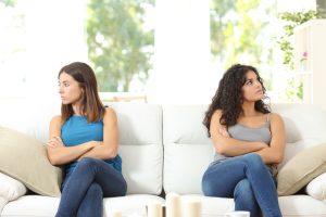 Sisters seated at opposite ends of sofa, arms crossed, looking away from each other