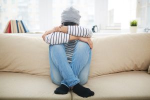 A young person sits on sofa with legs crossed, head on knees, face hidden