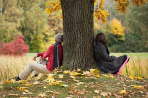 A couple sits looking away from each other on either side of a tree, with leaves scattered on the ground