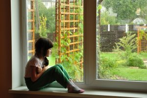 Child with braids sits on a windowsill and looks out into garden beyond