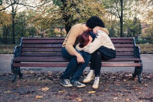 A sad young couple embraces and holds each other, faces hidden, on a park bench