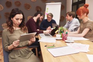 One person sits apart, looking down at tablet, in meeting of five people