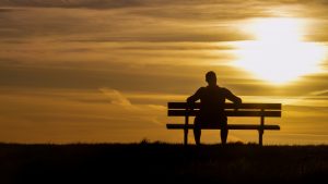 Rear view of silhouette of man sitting on bench watching the sun set in a cloudy gold sky