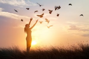 Person raises outstretched arms to sunset sky where flock of birds flies low