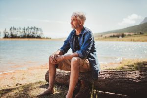 Senior citizen looks over water while relaxing on fallen log, calm expression on face