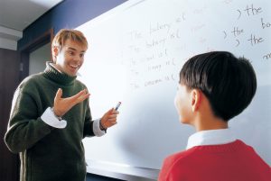 Young blond teacher stands at whiteboard and speaks to elementary-age student