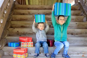 Two children sit next to a stack of presents. Both hold gifts on their heads. The younger child eyes the older child's larger gift. 