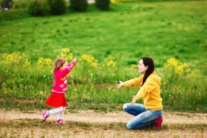 Young parent kneeling on path near grassy lawn holds arms open for toddler to run into