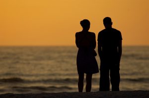 Rear view of silhouettes of a couple standing on shore and looking out to sea at sunset