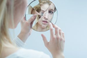 Person with long blonde hair looks into broken hand mirror to piece together reflection