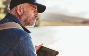 Mature individual wearing cap and overalls looks out over lake