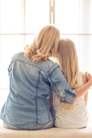 Rear view of adult with long blonde hair seated on sofa, arm around shoulder of teenager with long blonde hair