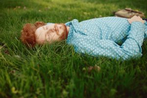 Young adult with red hair and beard, wearing light blue buttondown shirt, lies on grass with eyes closed