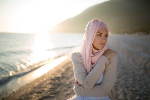 A woman who wears a hijab and has a serious expression hugs herself on the beach, looking off to the side