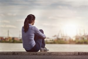 Rear view of person sitting on low wall under cloudy sky hugging knees and looking out over river