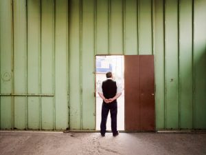 Zoomed-out rear view of person in suit vest and slacks with hands behind back standing in open doorway, looking outside