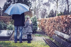 Tall adult in jeans and coat holds hand of child in dress coat with long hair as they stand at a gravestone on a rainy day