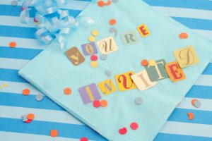 Cut-out letters on light blue napkin read "You're Invited." Confetti and ribbon surround the invitation.