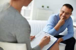 Seated mental health practitioner makes notes as seated person with half-smile describes concerns