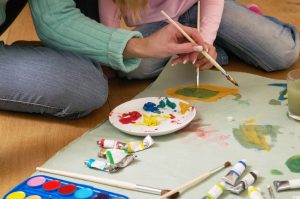 Focus on hands of mother in aqua sweater and child in pink sweater with blonde hair painting on large paper on floor