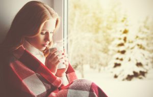 Person with long auburn hair sits in window, wrapped in red and white plaid blanket, inhaling steam from hot drink