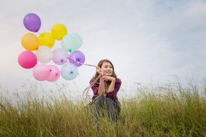 Young adult with long hair sits in tall grass, looking out into distance, holding a bunch of brightly colored balloons