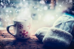 White mittents and red and white snowflake mug sit on wooden table with frosted window in background