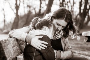 Black and white photo of adult with long hair putting arm around young teen with ponytail outside on bench