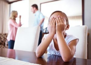 A little girl sits at the table and covers her face with her hands while her parents fight in the background