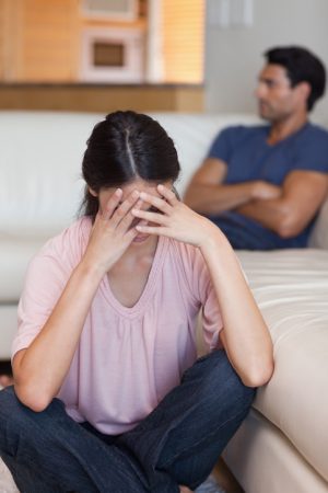 Person in background sits on sofa with arms crossed looking away from upset person in foreground who has head in hands