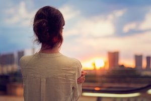 Rear view of person in casual blouse with hair in ponytail looking out over city at sunset in a blue sky with a few clouds