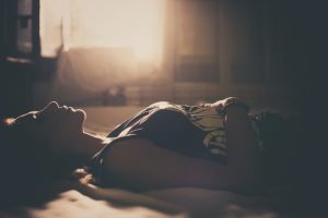 Person with short hair in tank top lies on bed, hands resting on stomach, eyes closer. Room is lit by sunset in sepia tones