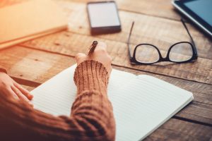 Close-up image of person's arm and hand, writing in open notebook at table. Glasses and smartphone sit nearby