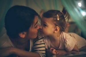 Young child and mother touch noses in dim room, gazing at each other affectionately