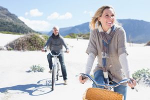 A middle-aged couple wearing warm clothes rides bikes along sandy beach, smiling and displaying enjoyment while looking off toward something in the distance