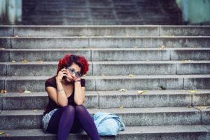 Young adult with curly hair dyed red wearing jean skirt and purple tights sits on stone steps outside, phone to ear, with serious expression