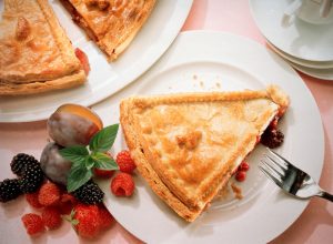 A slice of berry pie sits on a plate with a fork. There are berries to the side of the plate and a full pie nearby. 