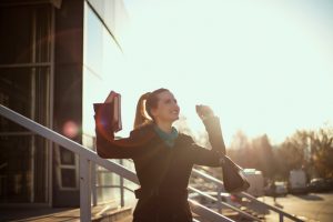 Person with ponytail wearing blazer and holding books raises fists in gesture of success on steps outside building