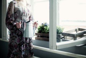 Person in long floral dress with head cropped out of photo stands near window planter while holding watering can tightly in both hands
