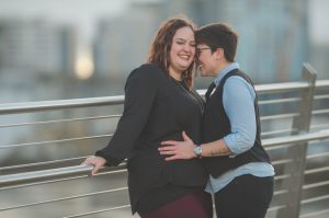 Young queer couple embrace happily while leanign on outdoor railing against bridge 