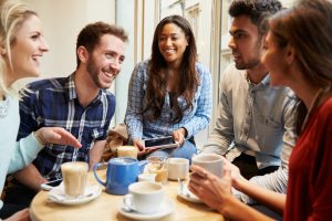 Five young people sit at table in coffee shop. One has digital device open. All are laughing and talking and looking at each other