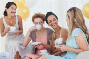 Small group of friends in dresses drink tea and laugh together on sofa at party