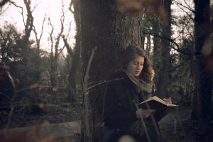 Twenty-something person with long hair, dressed in black and grey, sits by tree in woods and writes in journal