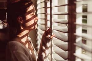 Person in sweater with pulled-back hair looks out through open blinds to reddish-tinted light