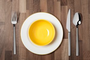 Empty yellow bowl on top of white plate with fork, knife, and spoon set alongside on top of wooden table