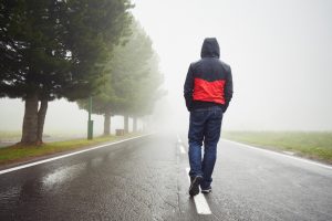 Rear view of person in black and red hooded sweatshirt walking alone down the middle of a road on a misty day