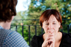 A person sits with hands clasped in front of face, thinking. Back of therapist's head is also visible